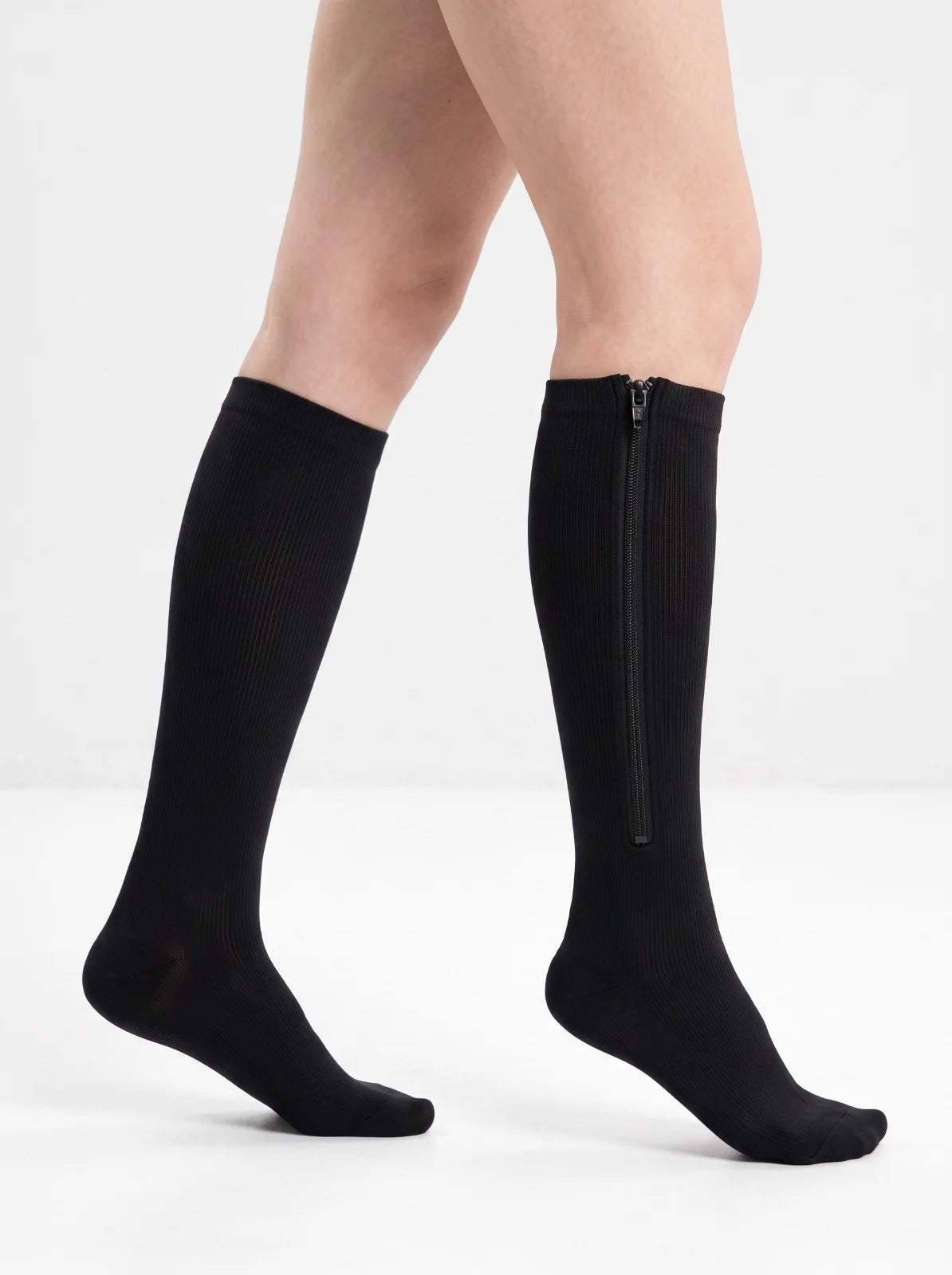 A pair of black knee-high compression socks with a side zipper, shown on a model's legs against a plain white studio background. 
